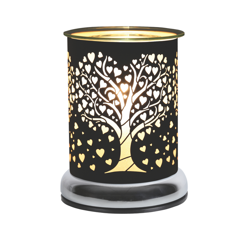 A cylindrical black electric wax melt warmer with glass dish emits warm light through a tree design with heart-shaped leaves cut out of the black metal casing, creating a cozy, romantic atmosphere.