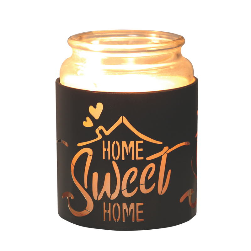 Lit candle in a glass jar with a black  holder, cut-out text "Home Sweet Home" and heart shapes, emits a warm glow, creating a cozy atmosphere.