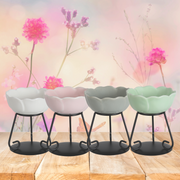 Four pastel-colored ceramic wax melt warmers on black metal stands, aligned on a wooden surface. Background features pink flowers and pastel tones, creating a serene, floral ambiance.