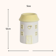 A ceramic house-shaped wax melt warmer with a soft yellow roof and windows. It measures 12cm tall, creating a cozy and whimsical vibe. Arrows indicate size.