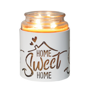 Lit candle in a glass jar with a white holder, cut-out text "Home Sweet Home" and heart shapes, emits a warm glow, creating a cozy atmosphere.