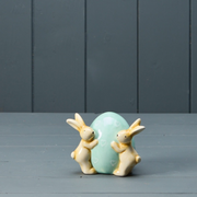 Two ceramic bunnies hug a large, pastel blue egg adorned with small heart shapes. They're set against a muted gray, wooden backdrop, conveying a whimsical and serene mood.