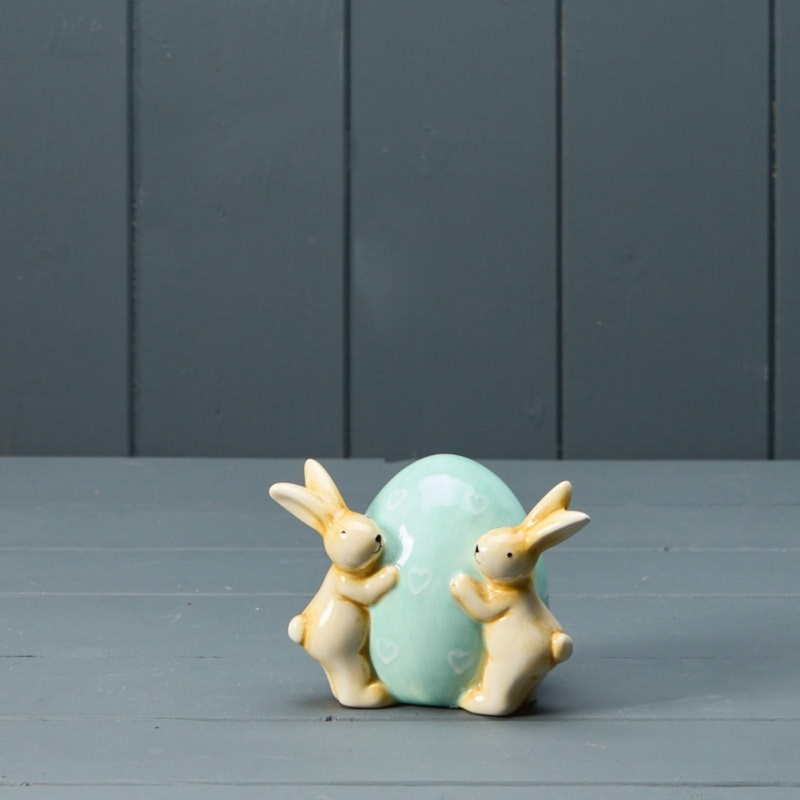 Two ceramic bunnies hug a large, pastel blue egg adorned with small heart shapes. They're set against a muted gray, wooden backdrop, conveying a whimsical and serene mood.