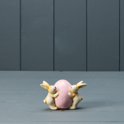 Two ceramic bunnies hug a large, pastel pink egg adorned with small heart shapes. They're set against a muted gray, wooden backdrop, conveying a whimsical and serene mood.