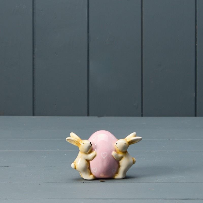Two ceramic bunnies hug a large, pastel pink egg adorned with small heart shapes. They're set against a muted gray, wooden backdrop, conveying a whimsical and serene mood.
