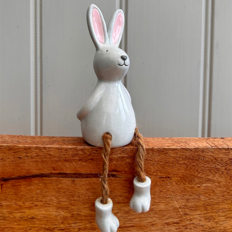 Ceramic bunny with pink ears sits on a wooden ledge. It has a cheerful expression and dangling rope legs with white ceramic feet, creating a playful tone.