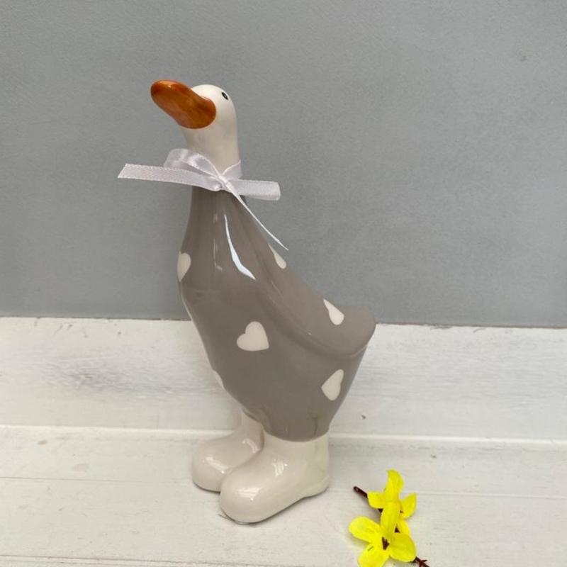 Ceramic duck ornament with a grey body, white heart patterns, and an orange beak. It wears a white ribbon around its neck, evoking a charming and whimsical tone.
