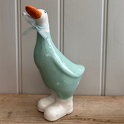 Ceramic duck figurine with a teal blue body, white polka dots, orange beak, light blue ribbon, and white boots, against a wooden backdrop. Playful tone.