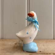 A ceramic duck figurine with blue polka dots and shoes, adorned with a blue ribbon around its neck. It stands on a wooden surface, exuding a whimsical charm.