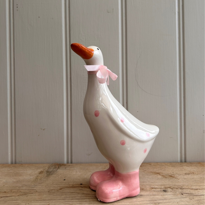 Ceramic duck with pink polka dots and bow, wearing pink boots, stands on wooden surface. Background is a gray paneled wall. Whimsical tone.