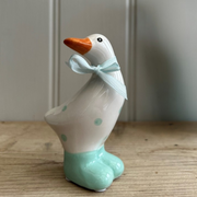 Ceramic duck figurine with a teal blue bow around its neck. It has a white body with teal blue spots and feet, set against a wooden surface.