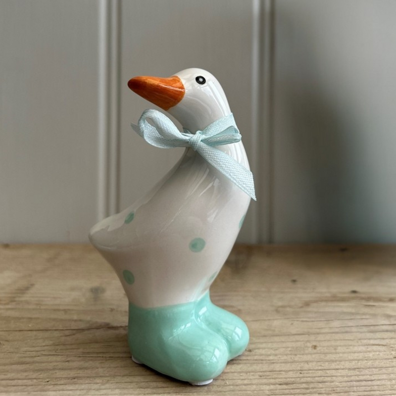 Ceramic duck figurine with a teal blue bow around its neck. It has a white body with teal blue spots and feet, set against a wooden surface.