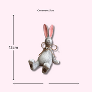A whimsical Laidback Sitting Bunny Rabbit Ornament figurine, Grey with pink ears and a twine bow sits playfully on a wooden surface, conveying a cheerful and rustic vibe.