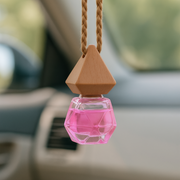 Hanging car air freshener with a geometric wooden cap and pink glass bottle, suspended in a car interior. The tone is calming and fresh.