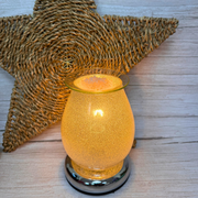 Glowing electric wax melt warmer on a wooden surface with a woven star backdrop. Warm ambiance with a metallic base, conveying cozy and rustic vibes.