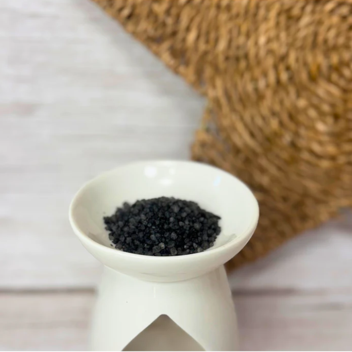 A white ceramic wax melt warmer holds grey fragrance scent sizzlers. In the background, a textured brown woven surface is partially visible, lending a rustic feel.