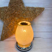 A glowing, electric wax melt warmer emits a warm light. It sits on a chrome base, with a woven star decoration in the background on a light wooden surface.