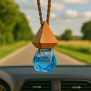 Hanging from a car's rearview mirror, a geometric blue glass car fragrance diffuser with a wooden cap reflects sunlight. A blurred road and green trees are in the background.