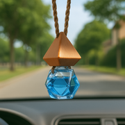 Hanging car fragrance diffuser. A blue geometric glass bottle, topped with a wooden cap, suspended against a blurred road and greenery background.