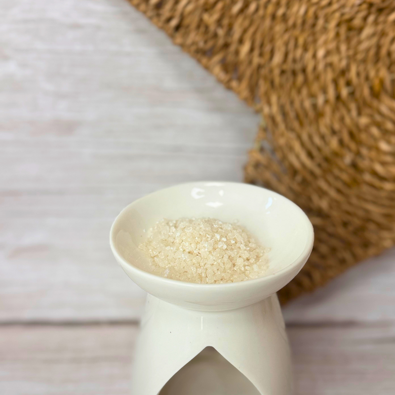 A white ceramic wax melt burner holds white fragrance crystals on a wooden surface, with a textured woven placemat partially visible in the background.