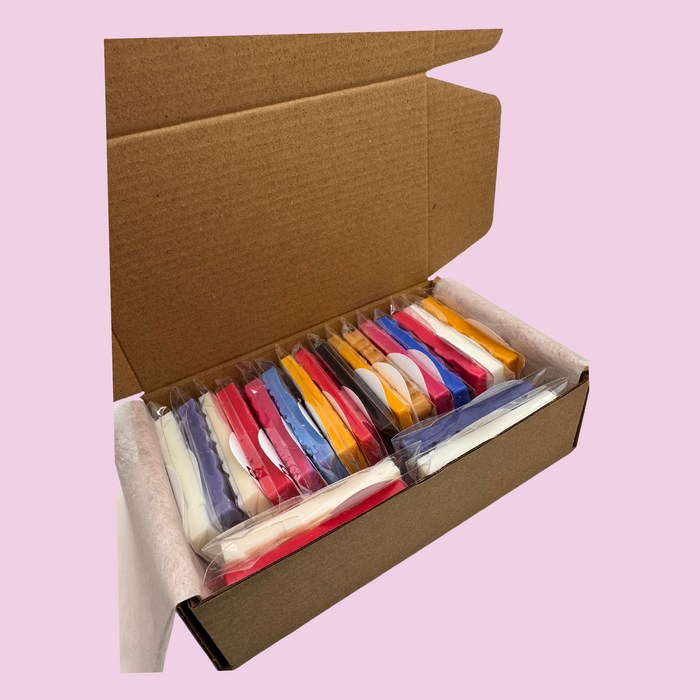 Open cardboard box on a pink background, containing neatly arranged colorful Wax Melt Mini Snap Bars in cellophane packets. Tones of red, blue, yellow, and white visible.