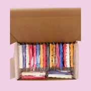 An open cardboard box reveals neatly stacked colorful wax melt mini snap bars. Each wax melt is packaged. Colours include red, blue, yellow, and pink on a soft pink background, conveying organization.