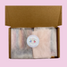 Open cardboard box on a pink background containing pastel-wrapped items with a round "Little House of Fragrance" sticker, conveying a delicate and inviting tone.