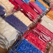 Colorful wax bars in various shapes, including purple and red with puzzle and geometric patterns, wrapped in clear plastic, displayed in rows.