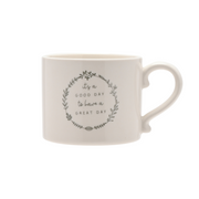 A white ceramic mug with a rounded handle features a motivational phrase: "It's a good day to have a great day" enclosed in a decorative leaf wreath.