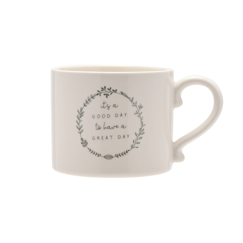 A white ceramic mug with a rounded handle features a motivational phrase: "It's a good day to have a great day" enclosed in a decorative leaf wreath.