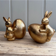 Two golden rabbit figurines are playfully hugging large golden eggs. Their pose suggests joy and whimsy, with a smooth and shiny metallic finish.