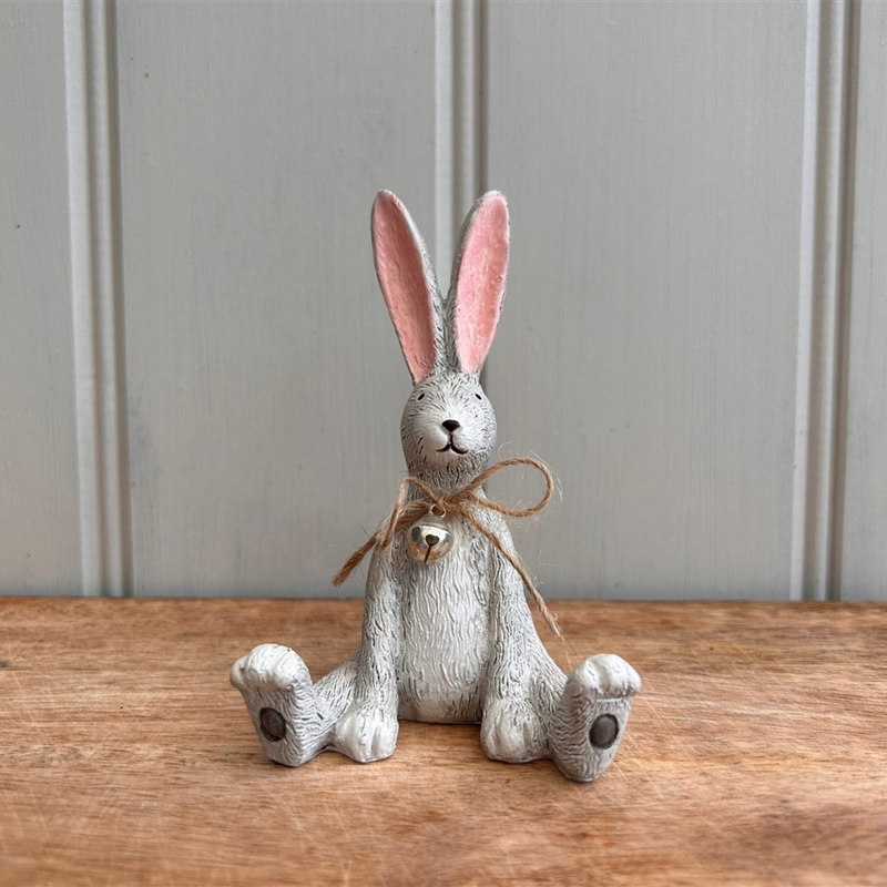A whimsical ceramic rabbit figurine with long ears and pink details sits on a wooden surface. It has a twine bow around its neck and an endearing expression.
