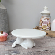 White ceramic cake stand with rabbit laying down holding the cake stand from above, on a wooden surface. Nearby, a cupcake and jar with star lid holding pastel cookies, creating a whimsical feel.
