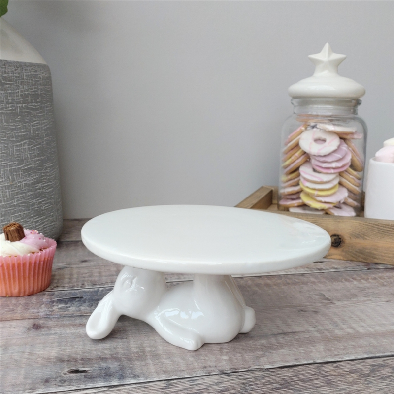 White ceramic cake stand with rabbit laying down holding the cake stand from above, on a wooden surface. Nearby, a cupcake and jar with star lid holding pastel cookies, creating a whimsical feel.
